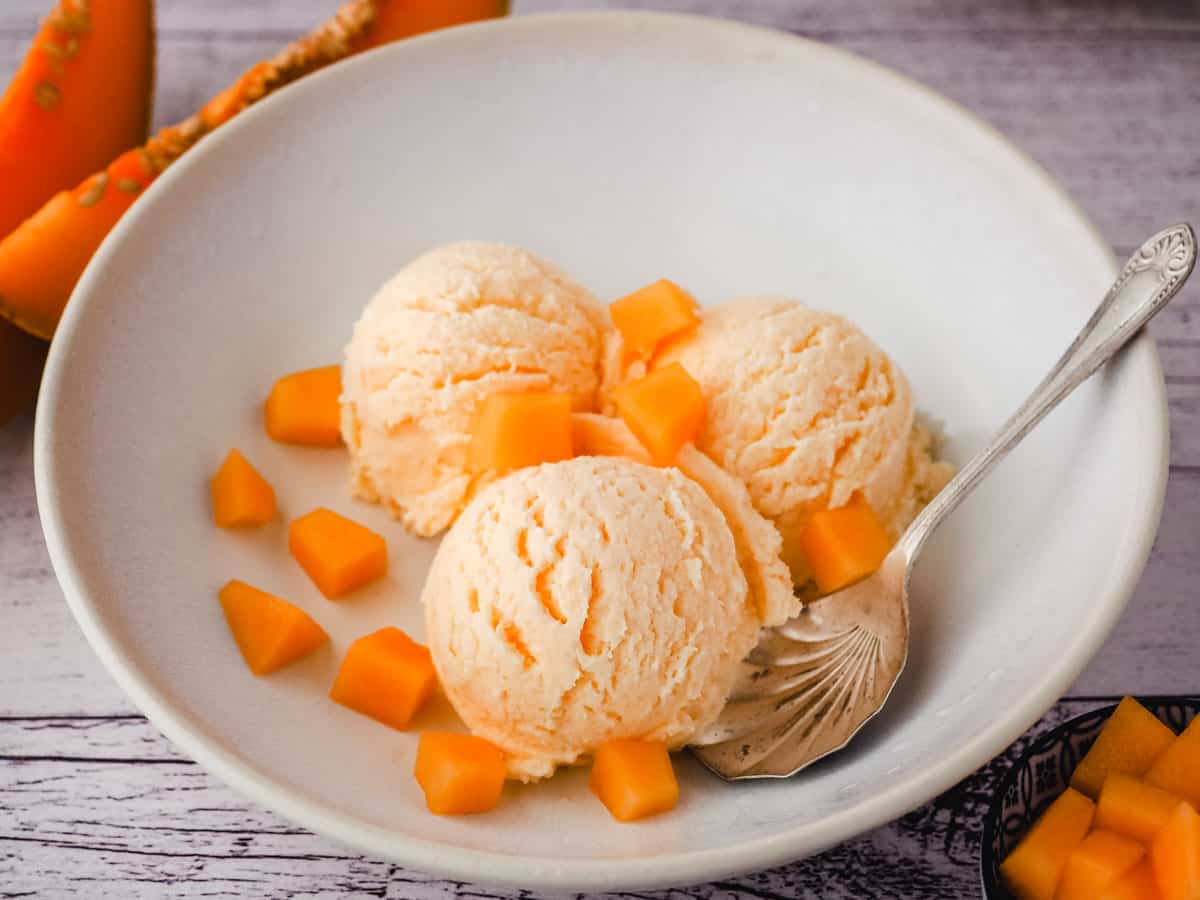 Three scoops of melon ice cream in a bowl, with a spoon and fresh melon pieces.