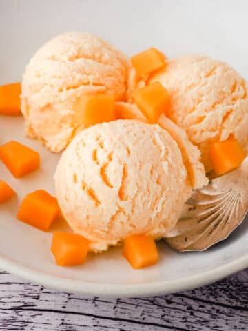 Three scoops of melon ice cream in a bowl, with a spoon and fresh melon pieces.
