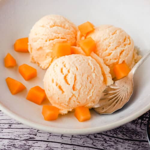 Three scoops of melon ice cream in a bowl, with a spoon and fresh melon pieces.