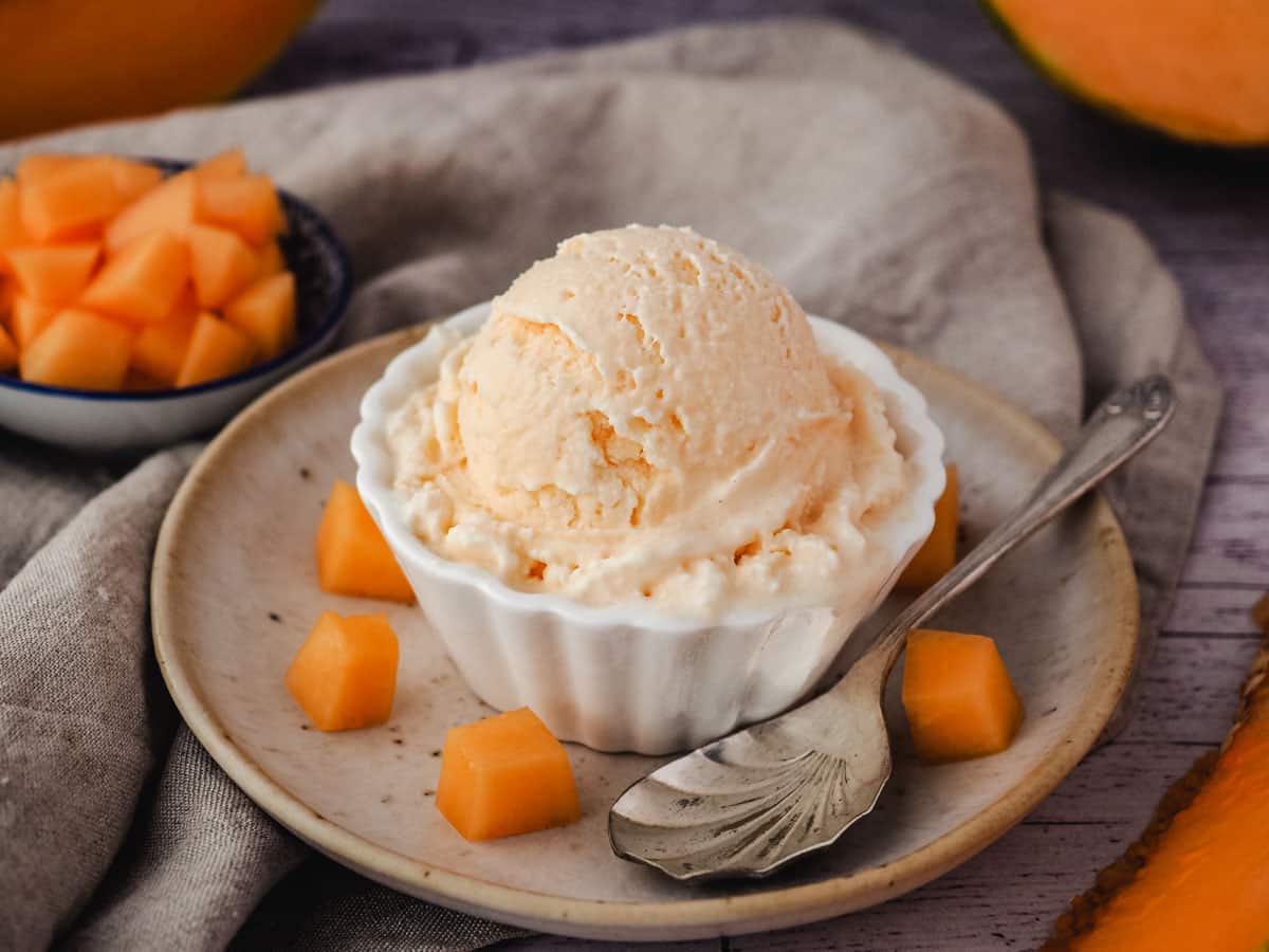 Single scoop of melon ice cream in a bowl, with a spoon and fresh melon on the side.