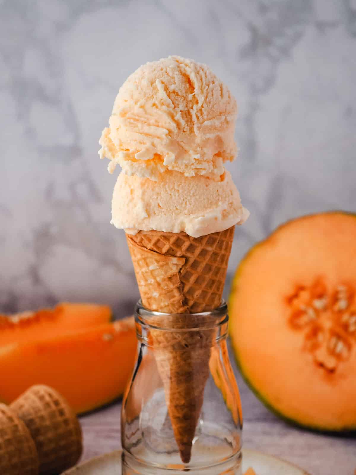 Two scoops of melon ice cream in a cone, with fresh melon in the background.