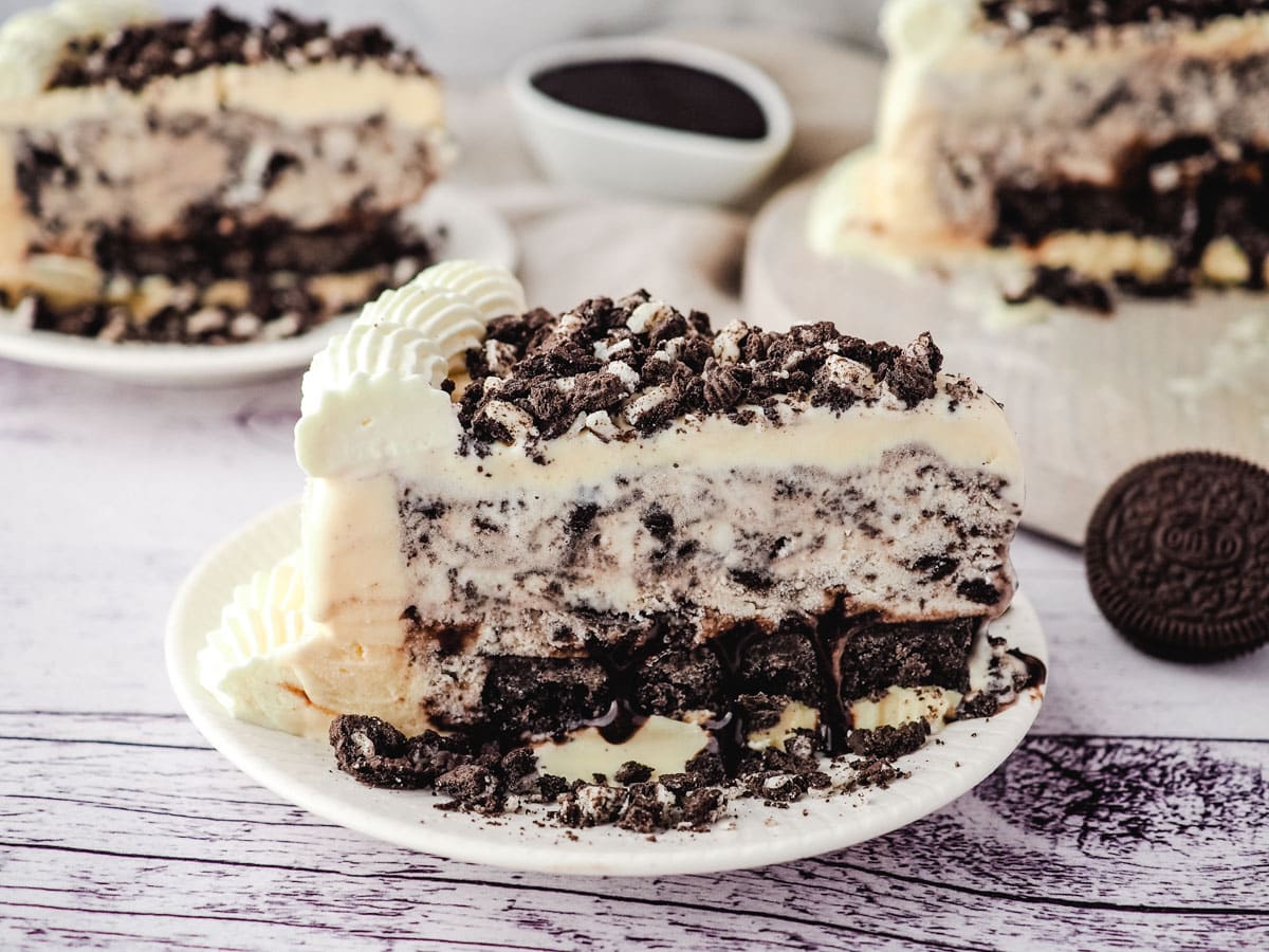Slice of Oreo ice cream cake on a plate, with another slice of cake, rest of cake and Oreos in the background.