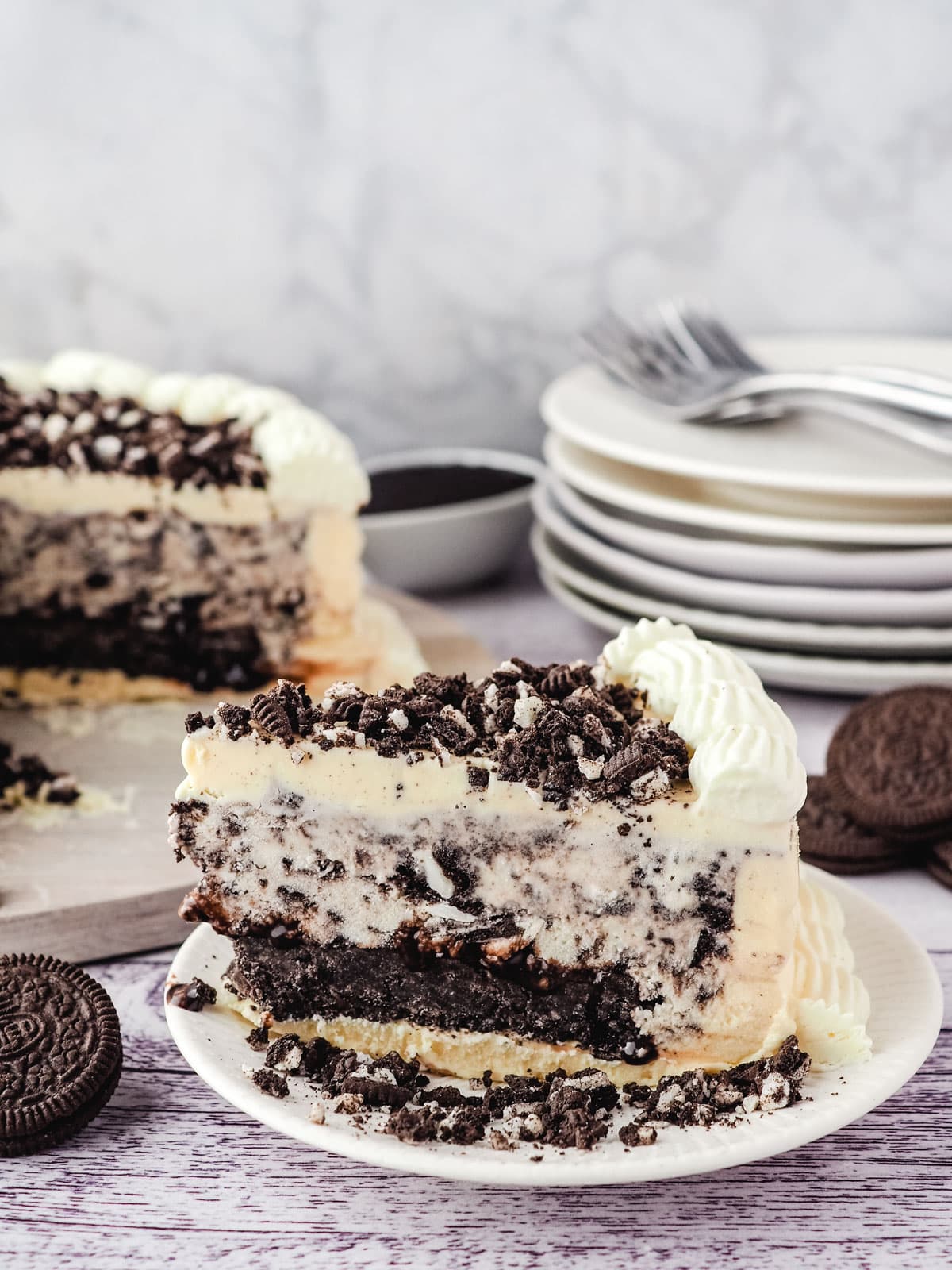 Slice of Oreo ice cream cake on a plate, with rest of cake and a stack of plate and forks in the background.