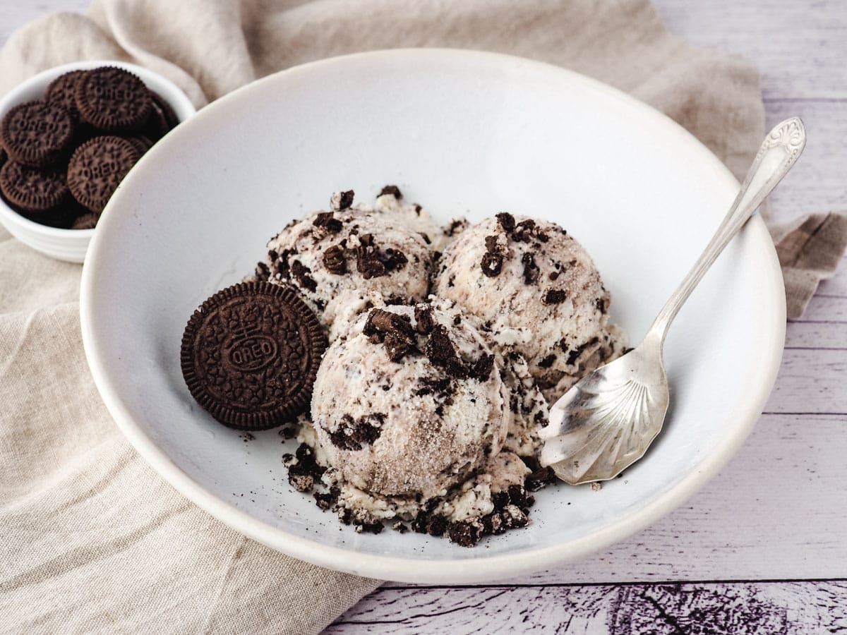 Three scoops of Oreo ice cream in a bowl with a spoon, and mini Oreos on the side.