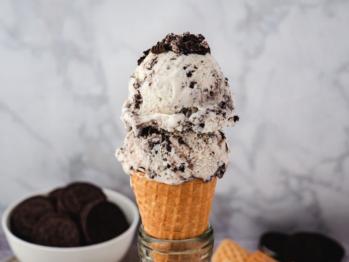 Two scoops of Oreo ice cream in a cone with Oreos in the background.