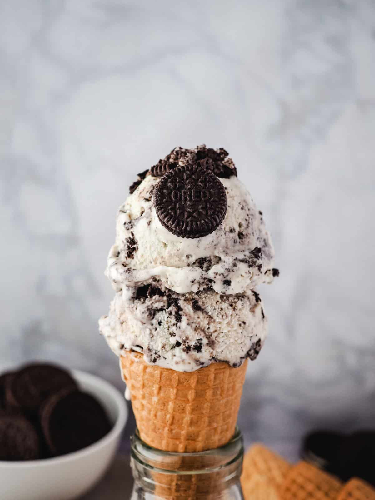 Two scoops of Oreo ice cream in a cone, garnished with a mini Oreo and Oreos in the background.
