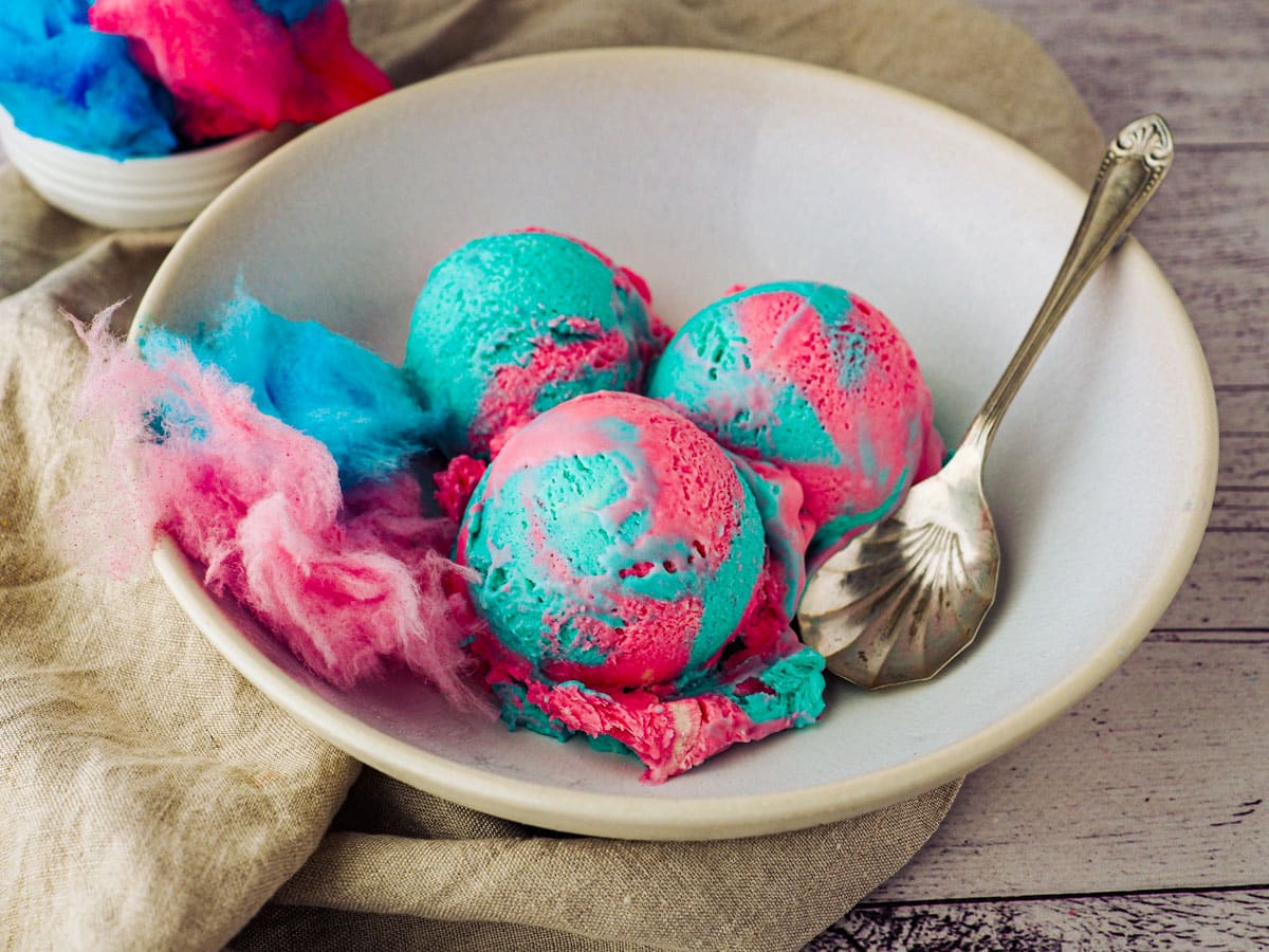 Scoops of cotton candy ice cream in a bowl with a spoon and pink and blue cotton candy.