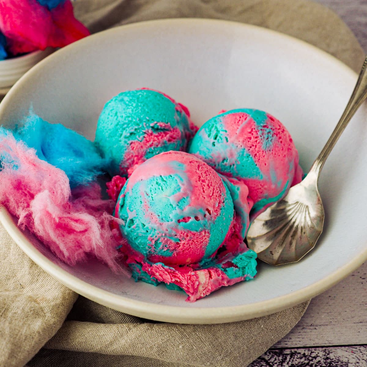 Scoops of cotton candy ice cream in a bowl with a spoon and pink and blue cotton candy.