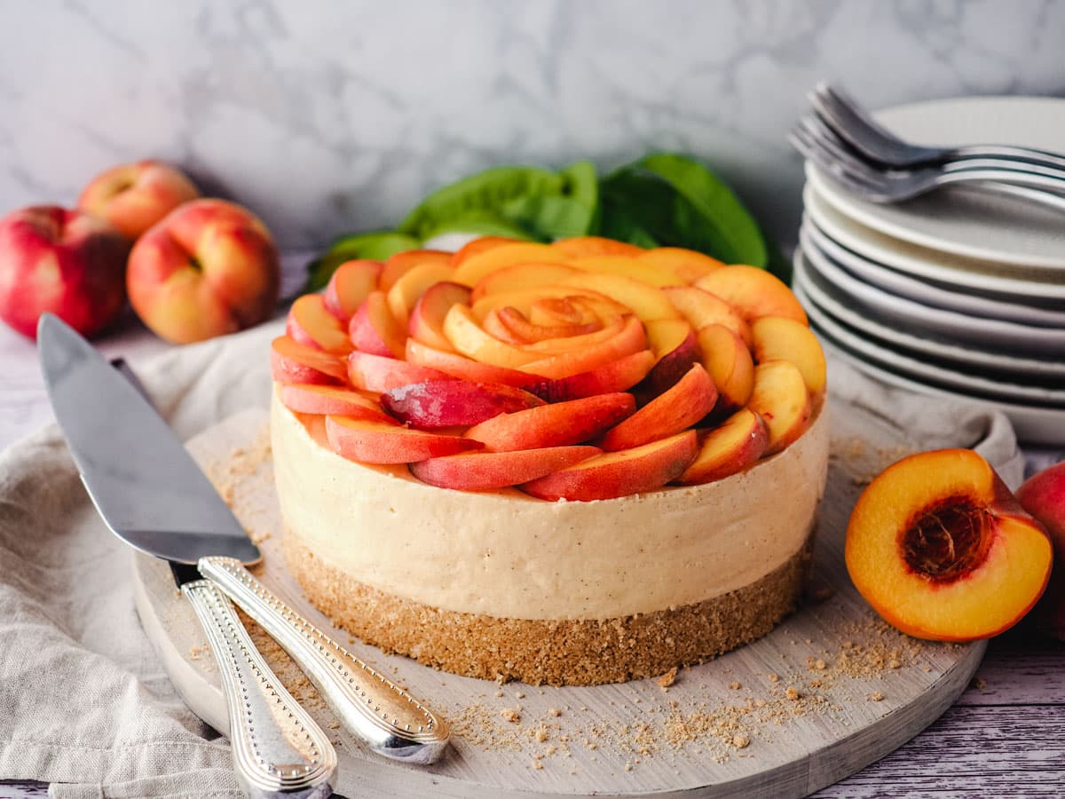 Peach cheesecake with fresh peach rose on a serving board, with a pile of plates and forks in the background.