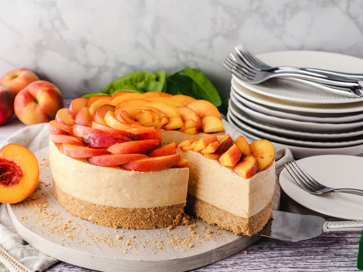 Removing a slice of peach cheesecake from the cake, with a pile of plates and forks in the background.