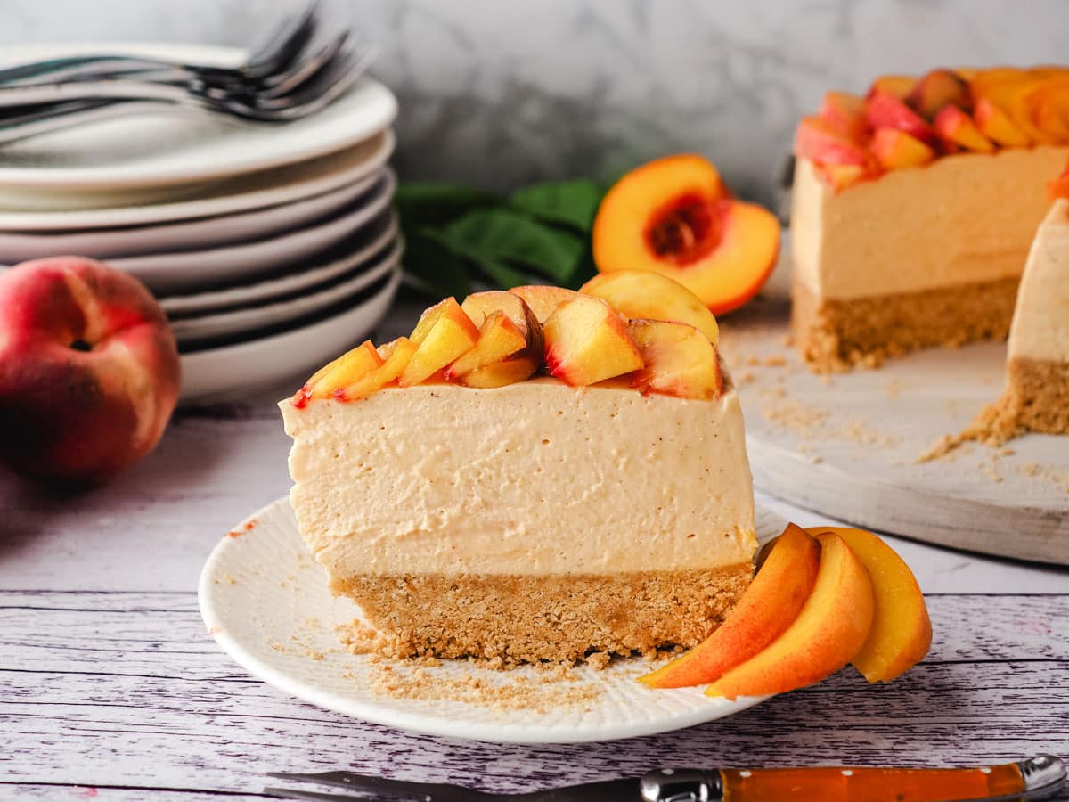 Slice of peach cheesecake with fresh peach rose on a plate with a fork, with rest of cake and pile of plates and forks in the background.