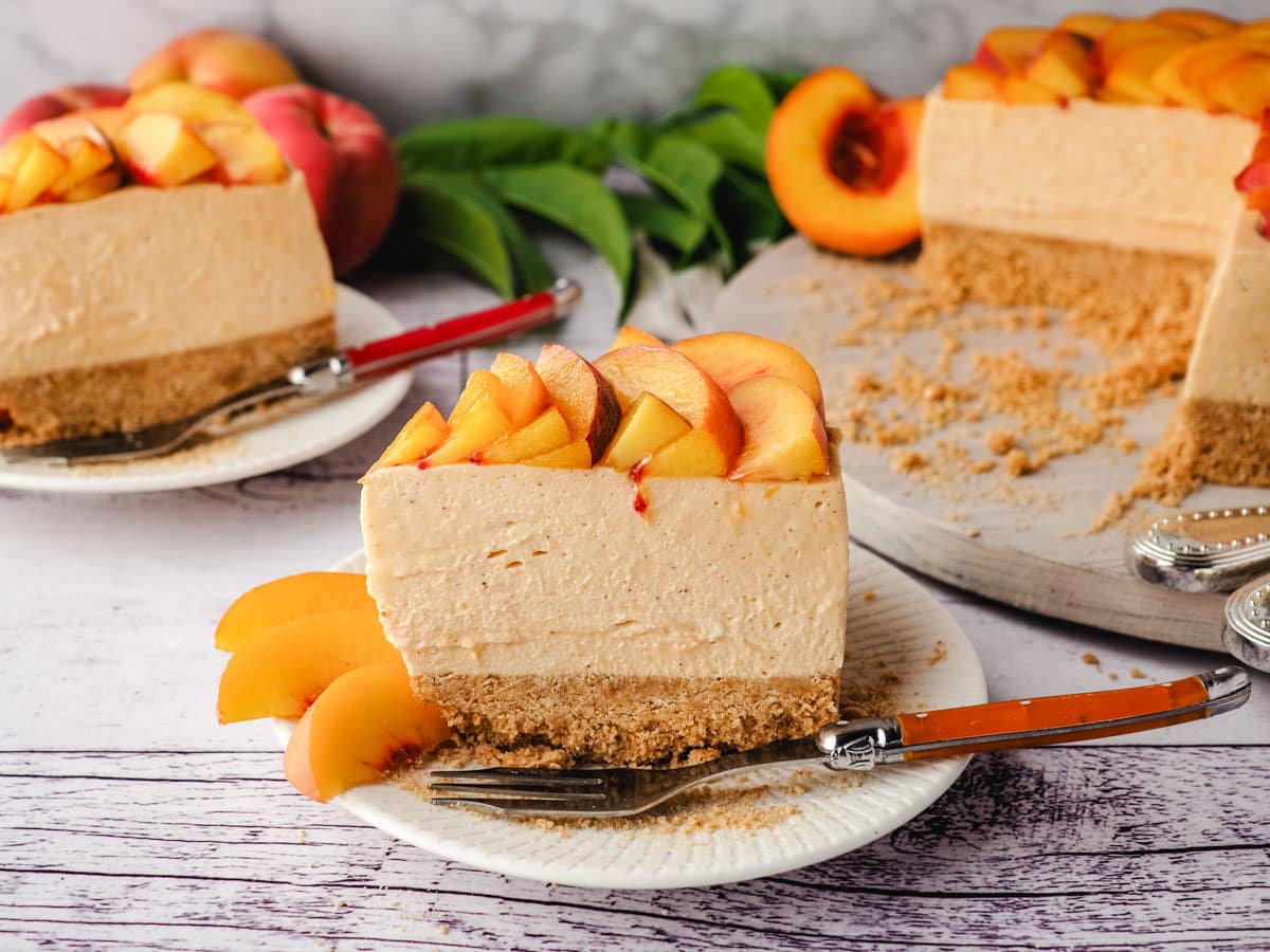 Slice of peach cheesecake with fresh peach rose on a plate with a fork, with rest of cake and another slide of cheesecake in the background.