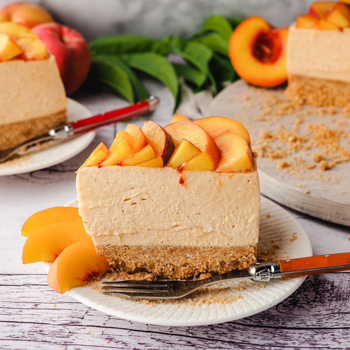 Slice of peach cheesecake with fresh peach rose on a plate with a fork, with rest of cake and another slide of cheesecake in the background.