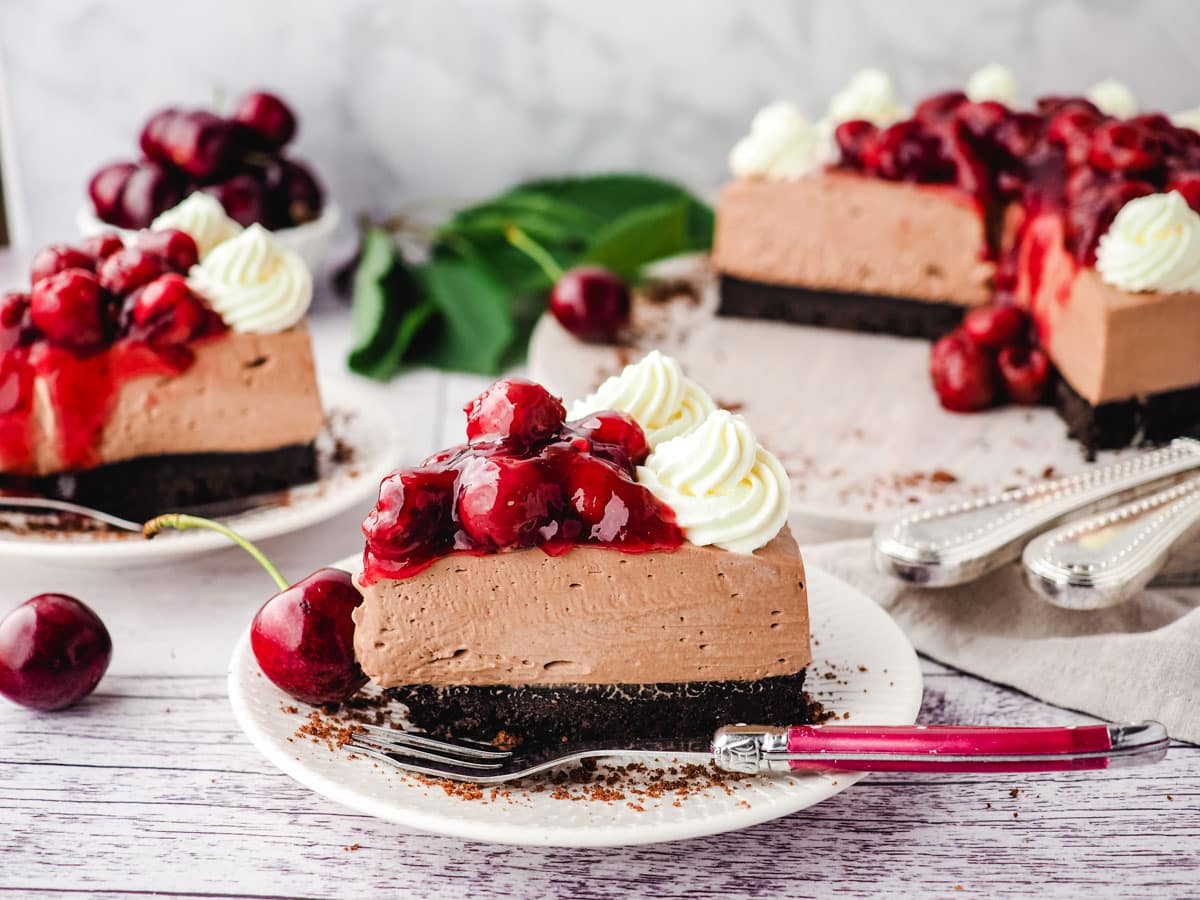 Slice of chocolate cherry cheesecake with a fork and rest of cake in the background.