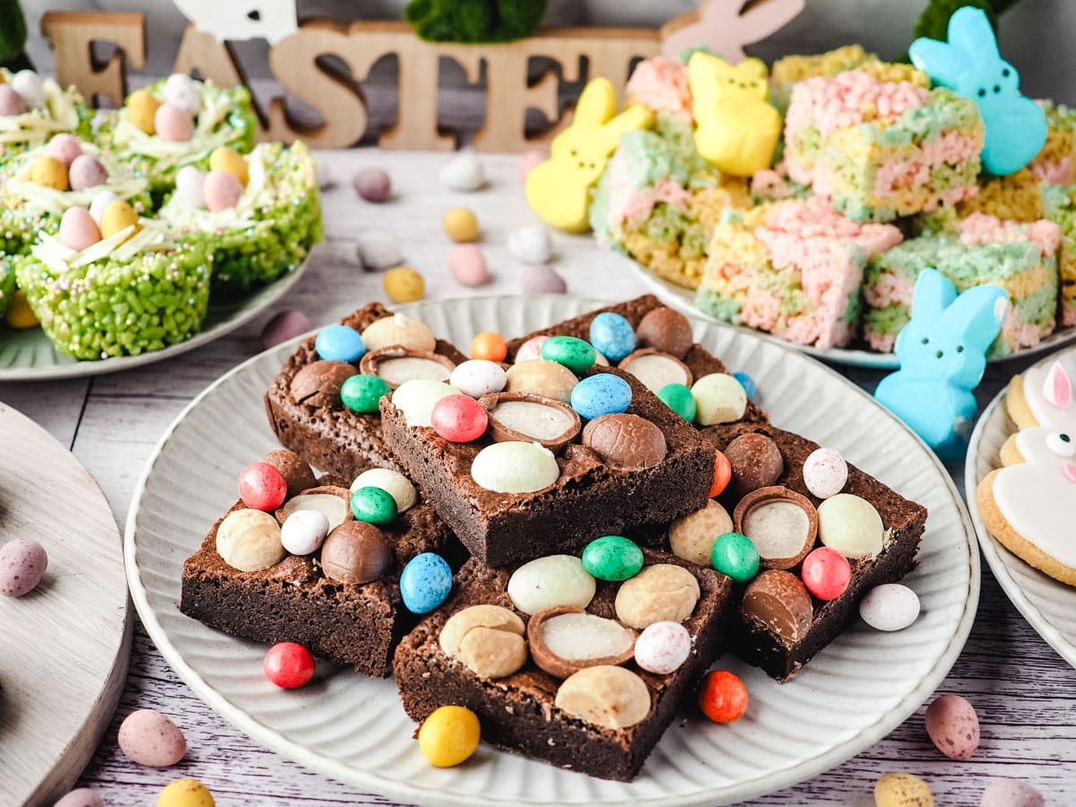 Easter brownies decorated with Easter eggs on a plate, on an Easter dessert table with bunny cookies, Easter rice krispie treats and peeps rice krispie treats in the background.