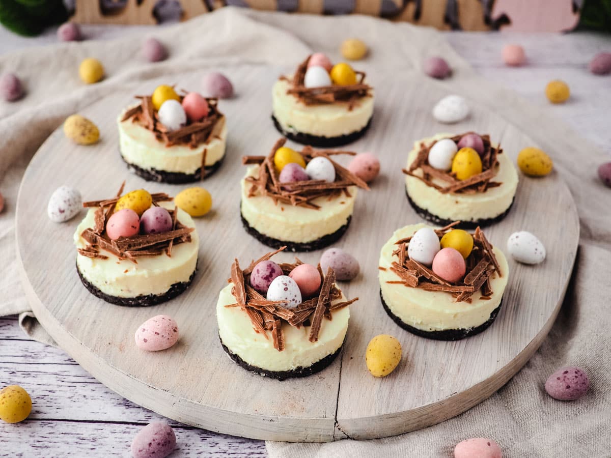 Mini Egg Cheesecakes 11 Mini egg cheesecakes on a serving board with Easter decorations in the background.