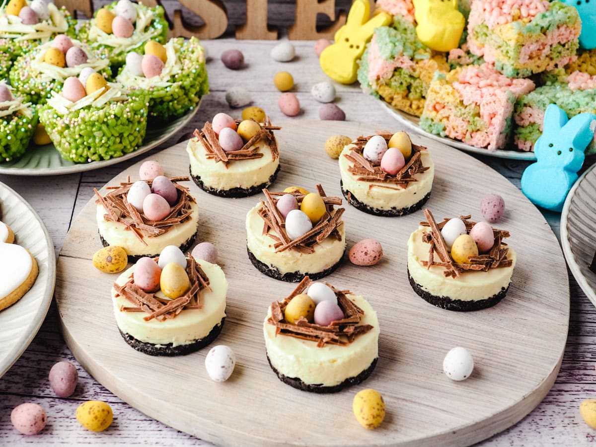 Mini egg cheesecakes on a serving board with Easter desserts and decorations in the background.