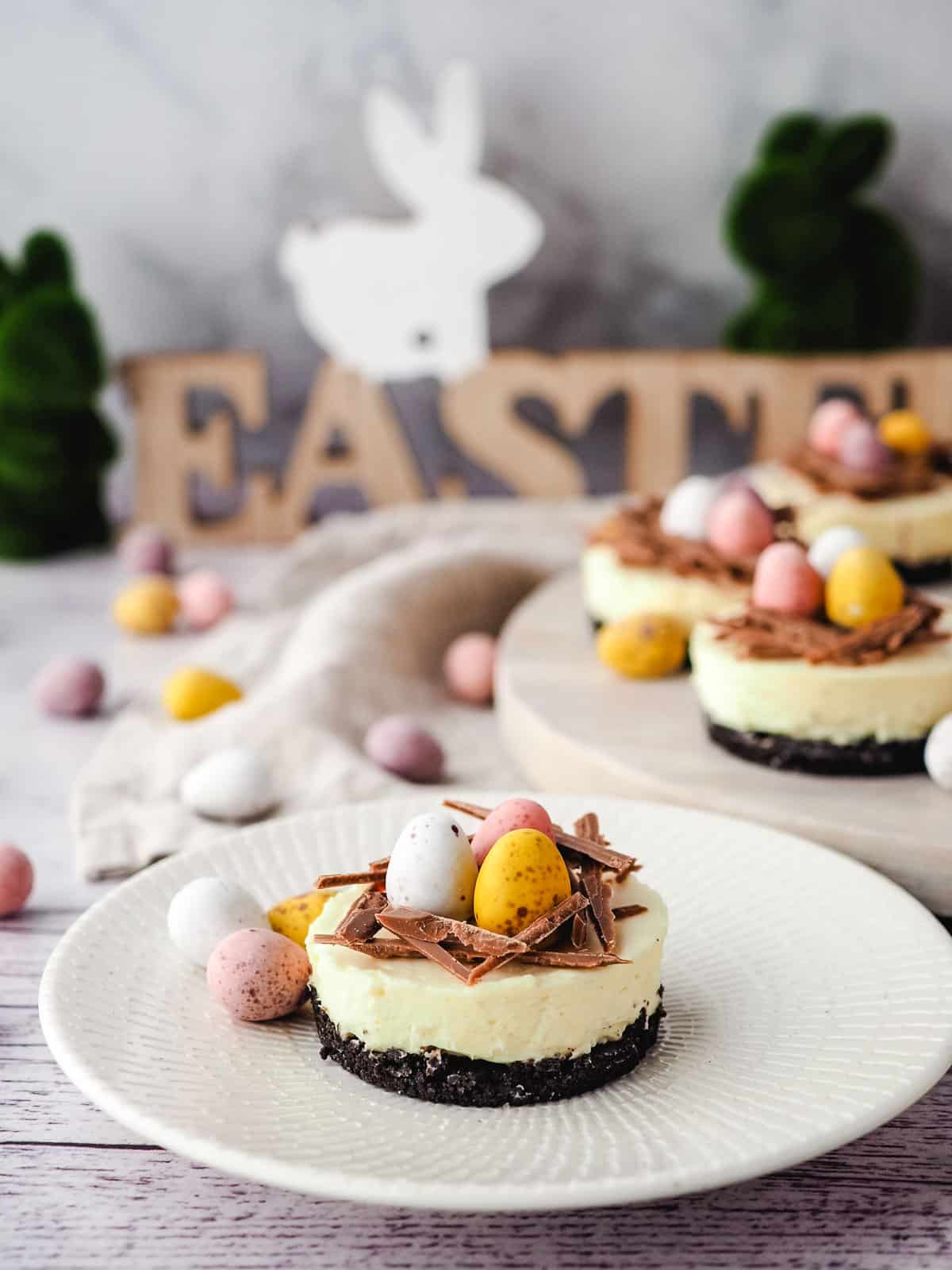 Mini egg cheesecake on a plate with mini eggs and rest of cheesecakes on a serving board in the background with Easter decorations.