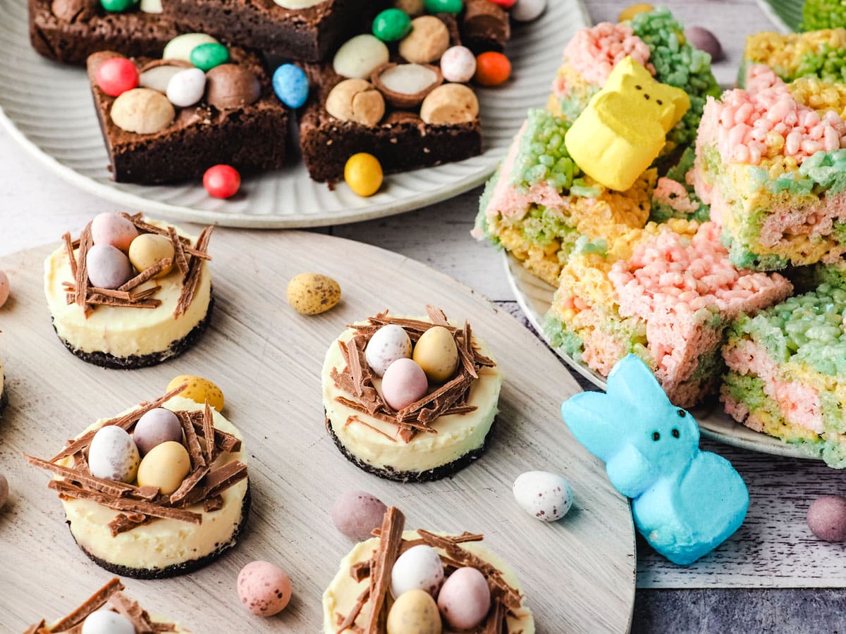 Mini Egg Cheesecakes 9 Mini egg cheesecakes on a serving board with Easter desserts and decorations in the background.