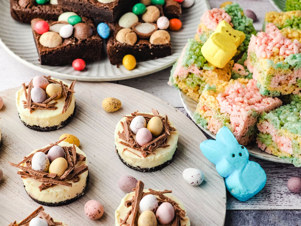 Mini egg cheesecakes on a serving board with Easter desserts and decorations in the background.