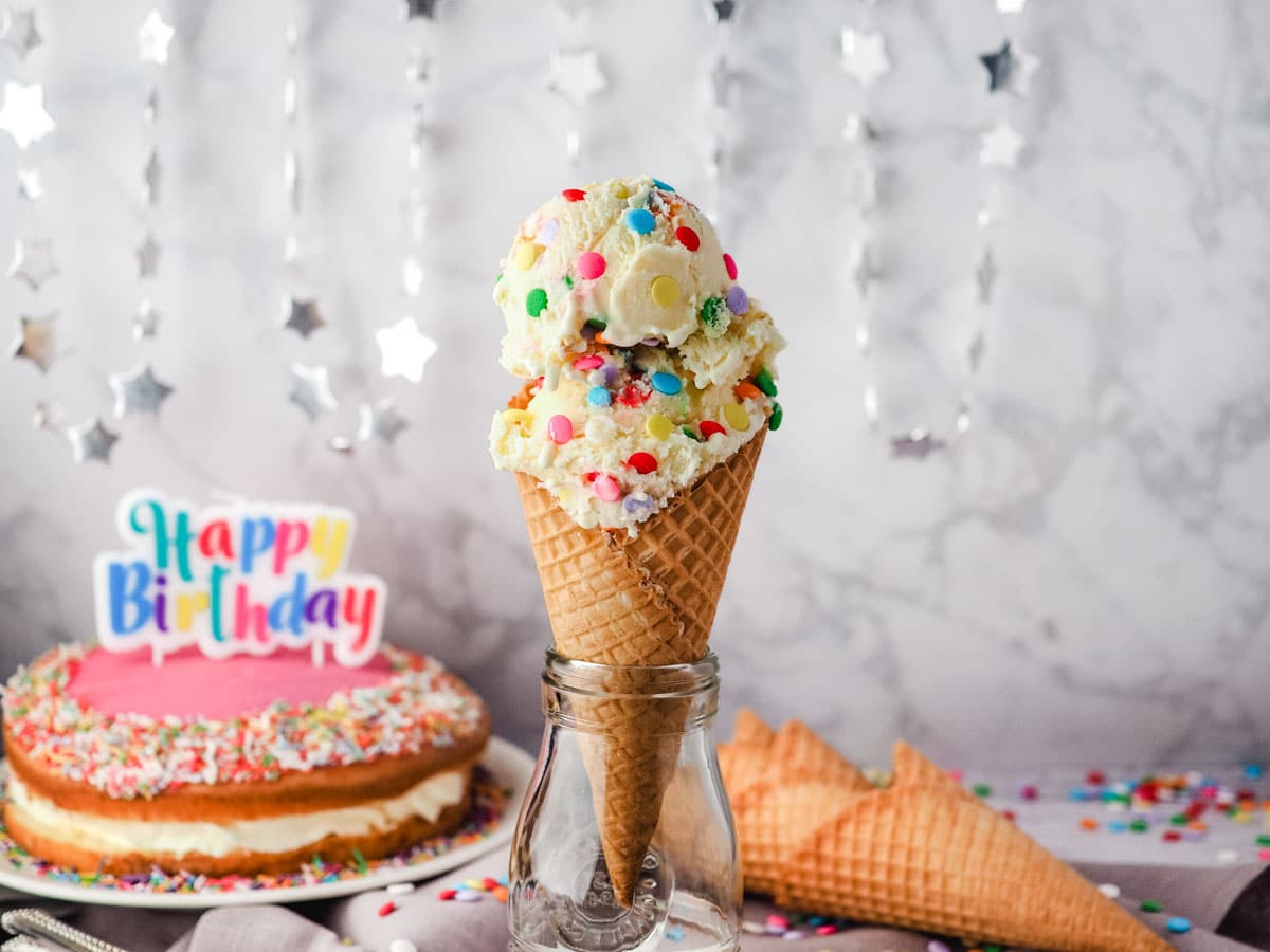 Two scoops of birthday cake ice cream in a cone with cones, sprinkles and a birthday cake in the background.