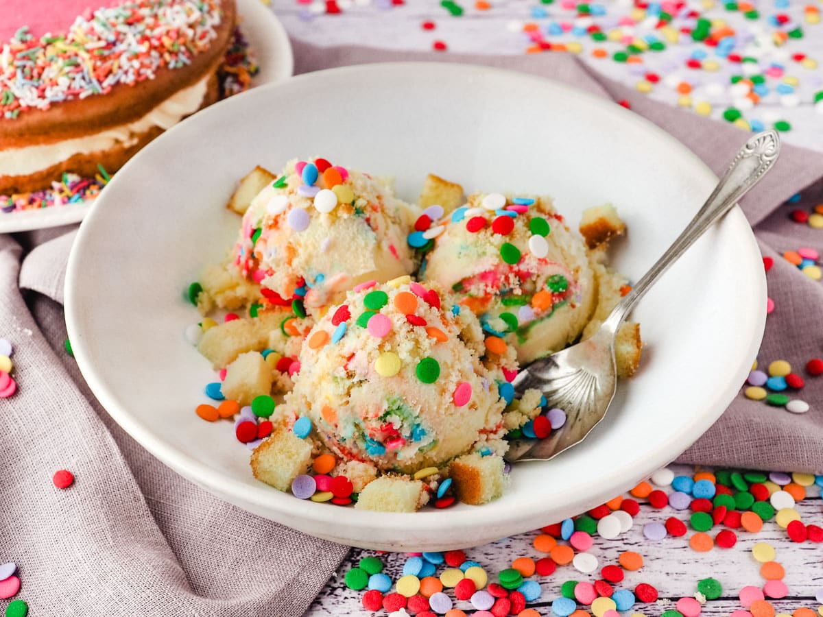 Scoops of birthday cake ice cream in a bowl with a spoon, surrounded by sprinkles, with a birthday cake in the background.