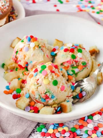 Scoops of birthday cake ice cream in a bowl with a spoon, surrounded by sprinkles.