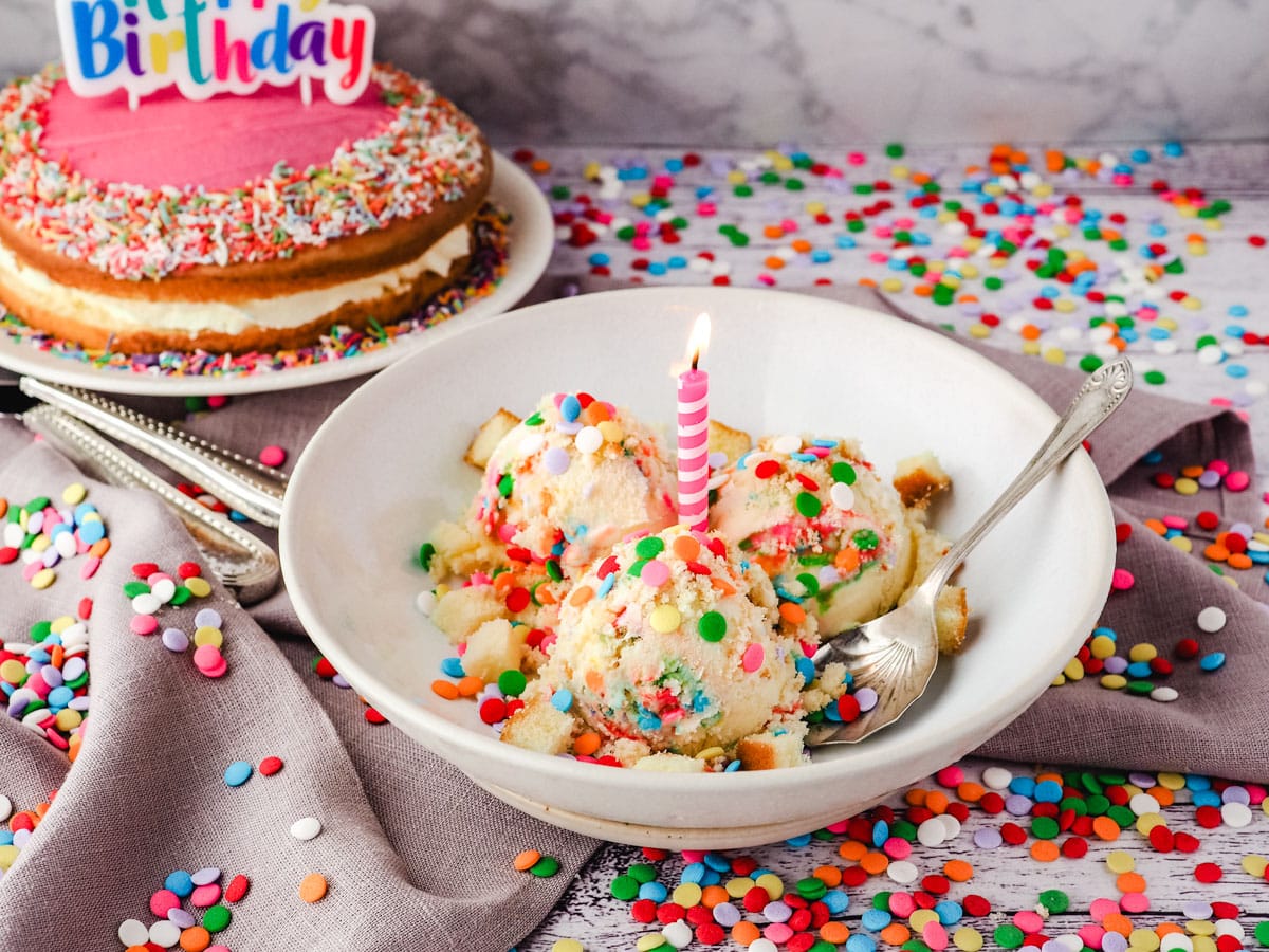 Scoops of birthday cake ice cream with a lite candle in a bowl with a spoon, surrounded by sprinkles, with a birthday cake in the background.