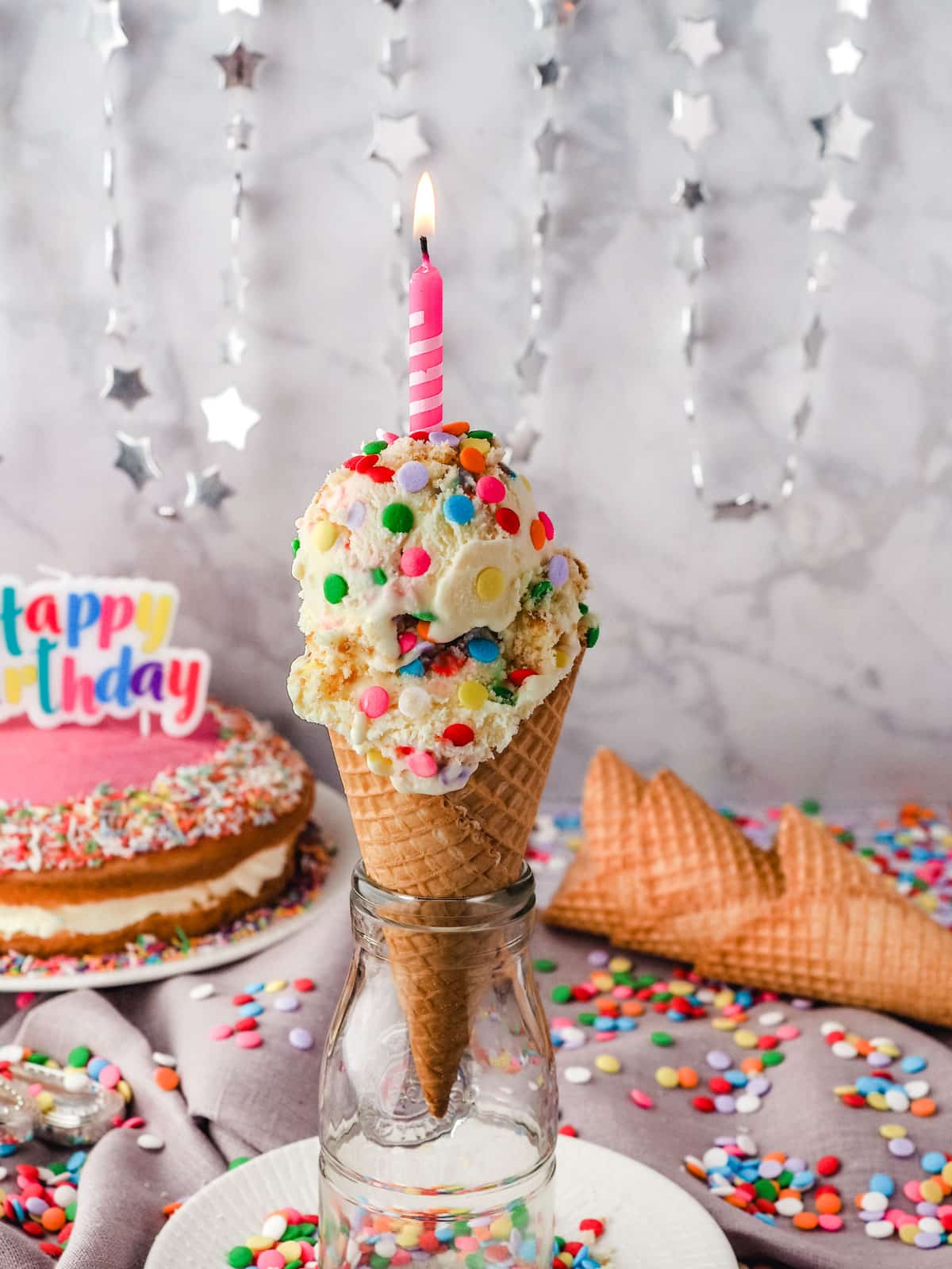 Two scoops of birthday cake ice cream in a cone with a lit birthday candle on top, with cones, sprinkles and a birthday cake in the background.