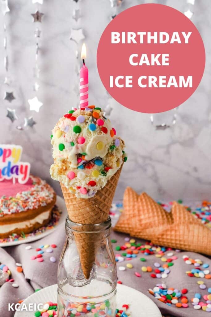 Two scoops of birthday cake ice cream in a cone with a lit birthday candle on top, with cones, sprinkles and a birthday cake in the background.