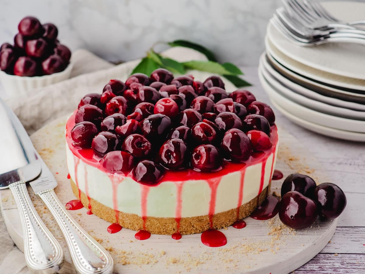 Cherry cheesecake on a serving platter with fresh cherries and serving ware on the side.