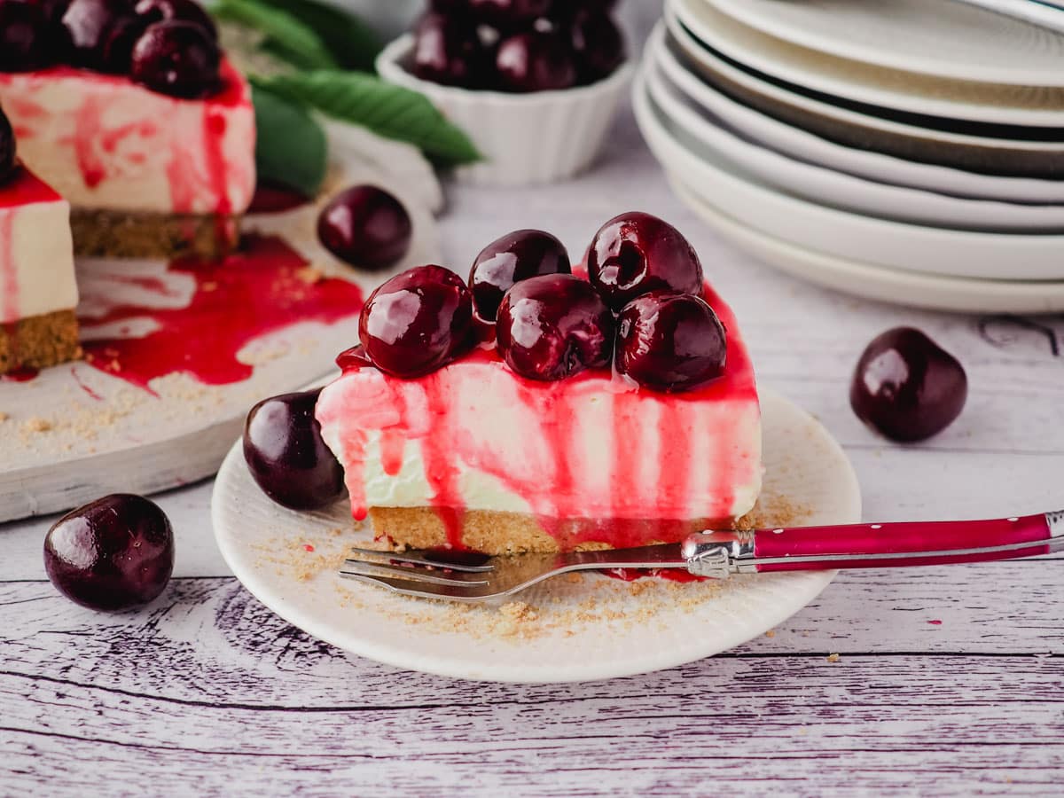Cherry Cheesecake 9 A slice of cherry cheesecake on a plate with a fork, with fresh cherries on the side and rest of cake and stack of plates in the background.