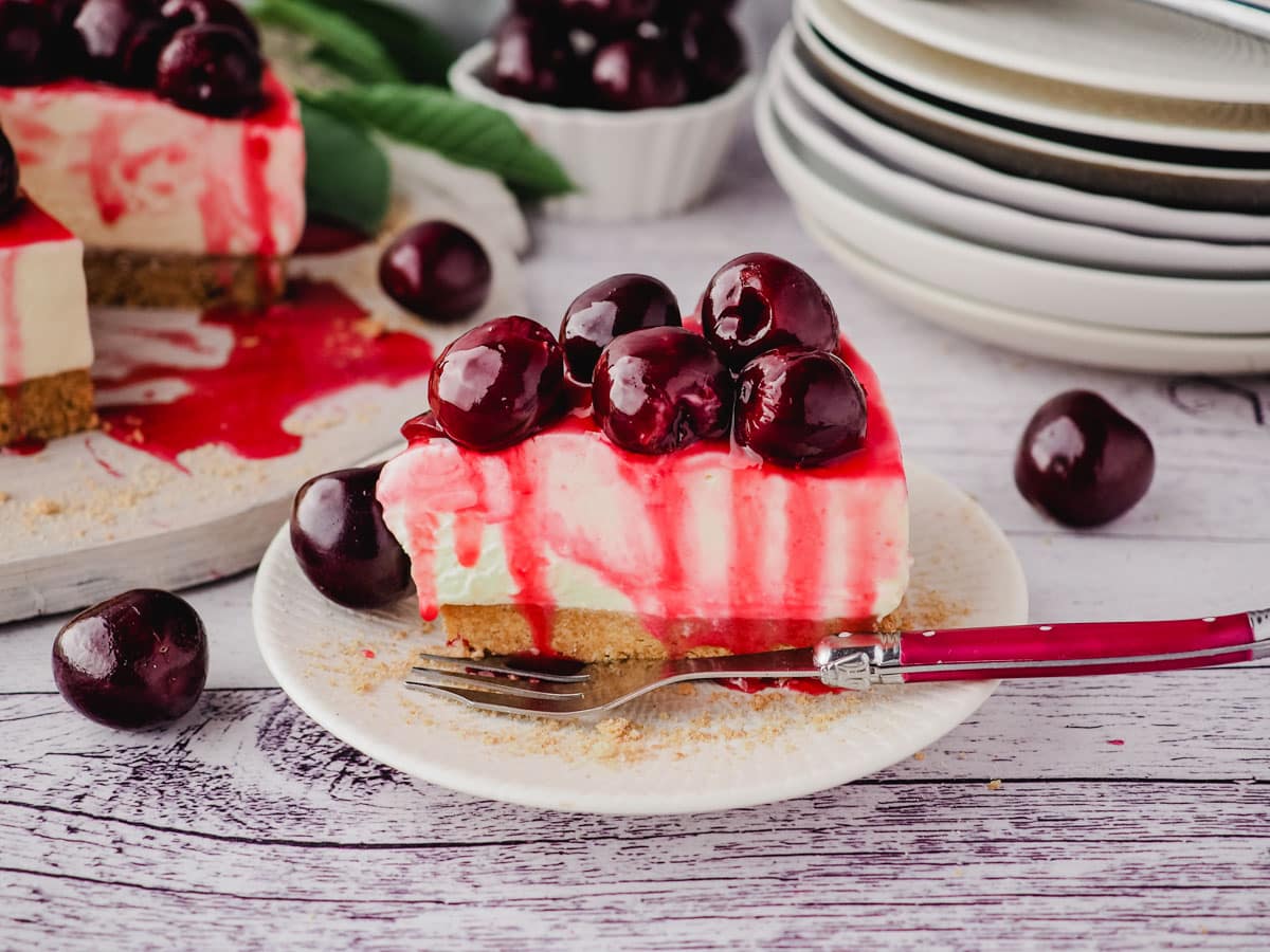 A slice of cherry cheesecake on a plate with a fork, with fresh cherries on the side and rest of cake and stack of plates in the background.