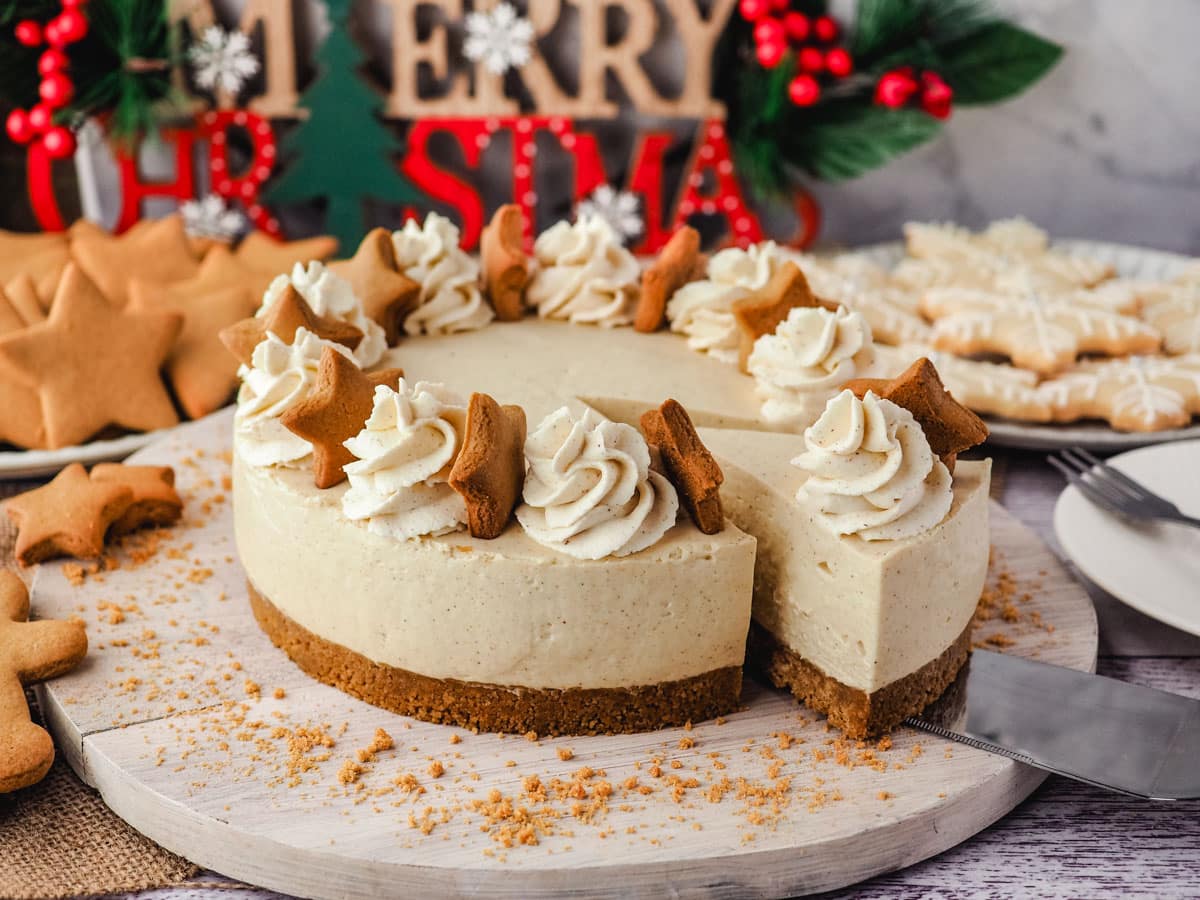 No Bake Cheesecake Recipes 10 Slicing a piece of gingerbread cheesecake on a serving board, surrounded by Christmas desserts and decorations.