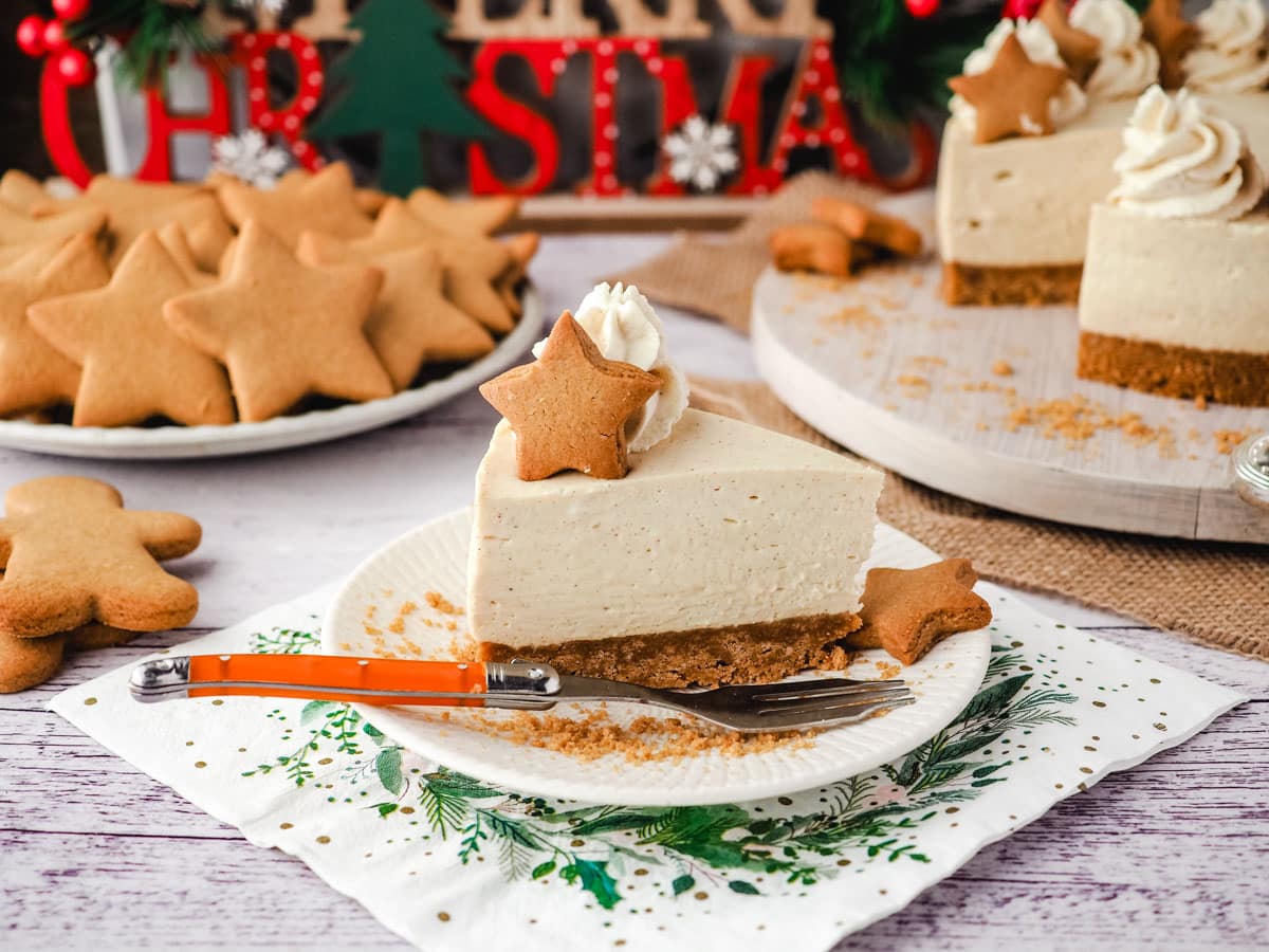 Slice of gingerbread cheesecake on a plate with a fork, surrounded by gingerbread cookies, rest of cheesecake and and Christmas decorations.