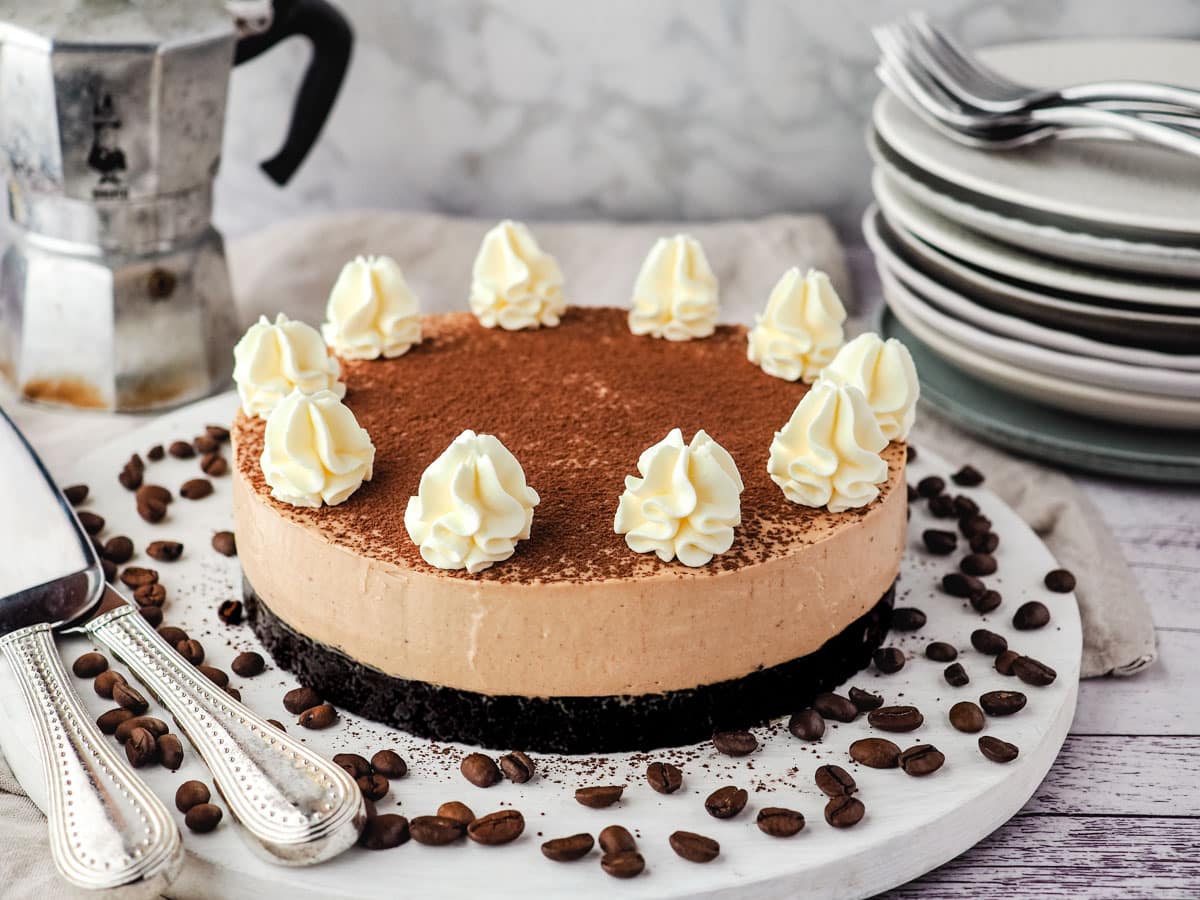 Coffee cheesecake on a serving board surrounded by coffee beans, with a stack of plates and forks and an espresso maker in the background.