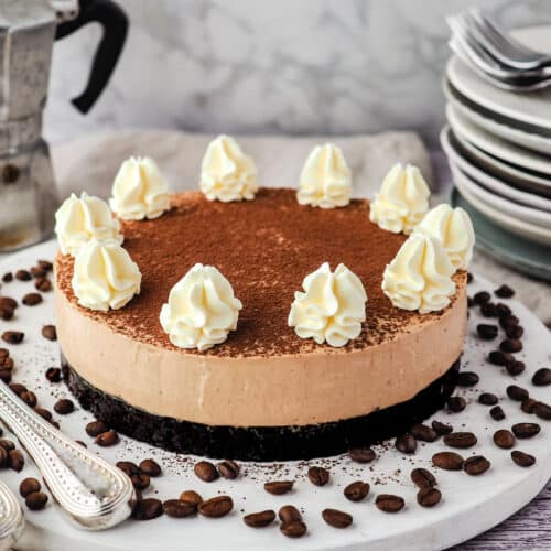 Coffee cheesecake on a serving board surrounded by coffee beans, with a stack of plates and forks and an espresso maker in the background.