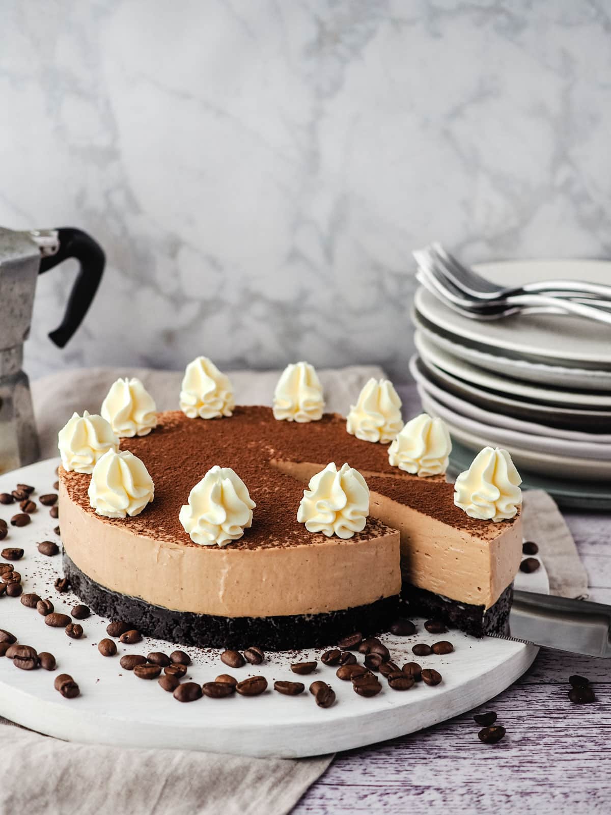 Coffee cheesecake with a slice being taking out of it, with a stack of plates and forks and an espresso maker in the background.