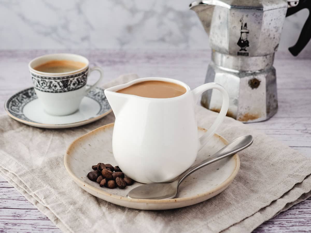 Coffee sauce in a serving jug with a spoon and coffee beans on the side, and cup of express and espresso pot in the background.