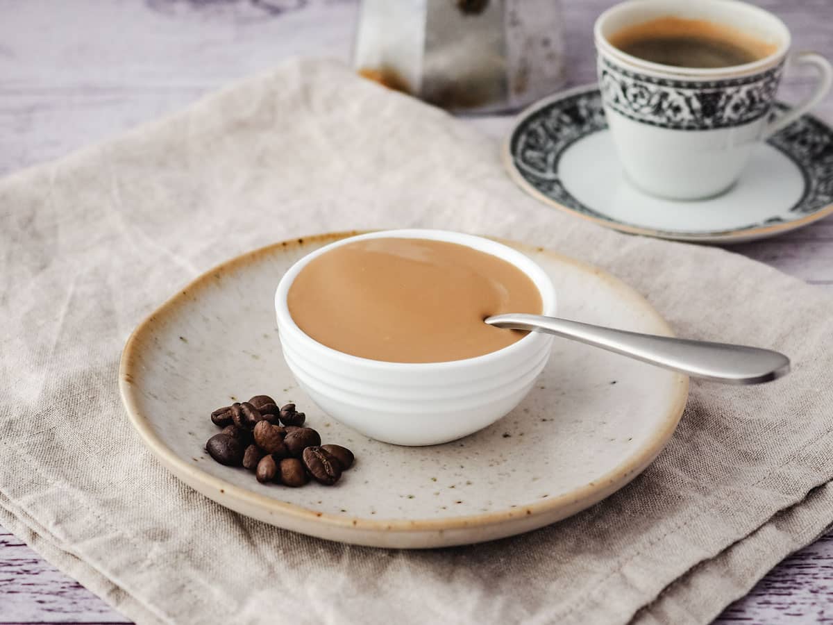 Coffee sauce in a serving bow with a spoon and coffee beans on the side, and cup of express and espresso pot in the background.