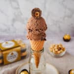 Scoops of Ferrero Rocher ice cream in a cone with a cut-open Ferrero on top, with Ferrero Rocher in the background.