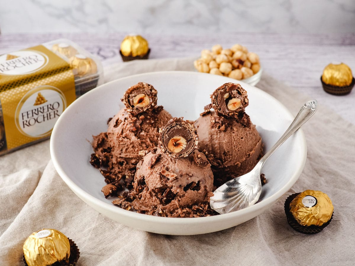 Three scoops of Ferrero Rocher ice cream with cut open Ferrero's on top, in a bowl, with Ferrero Rocher in the background.