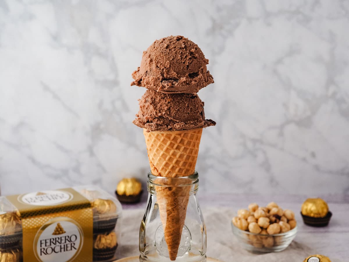 Scoops of Ferrero Rocher ice cream in a cone, with Ferrero Rocher in the background