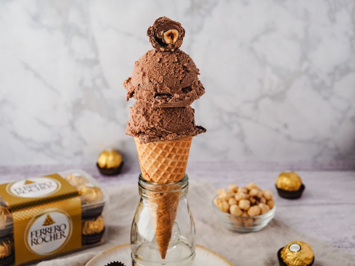 Scoops of Ferrero Rocher ice cream in a cone with a cut-open Ferrero on top, with Ferrero Rocher in the background
