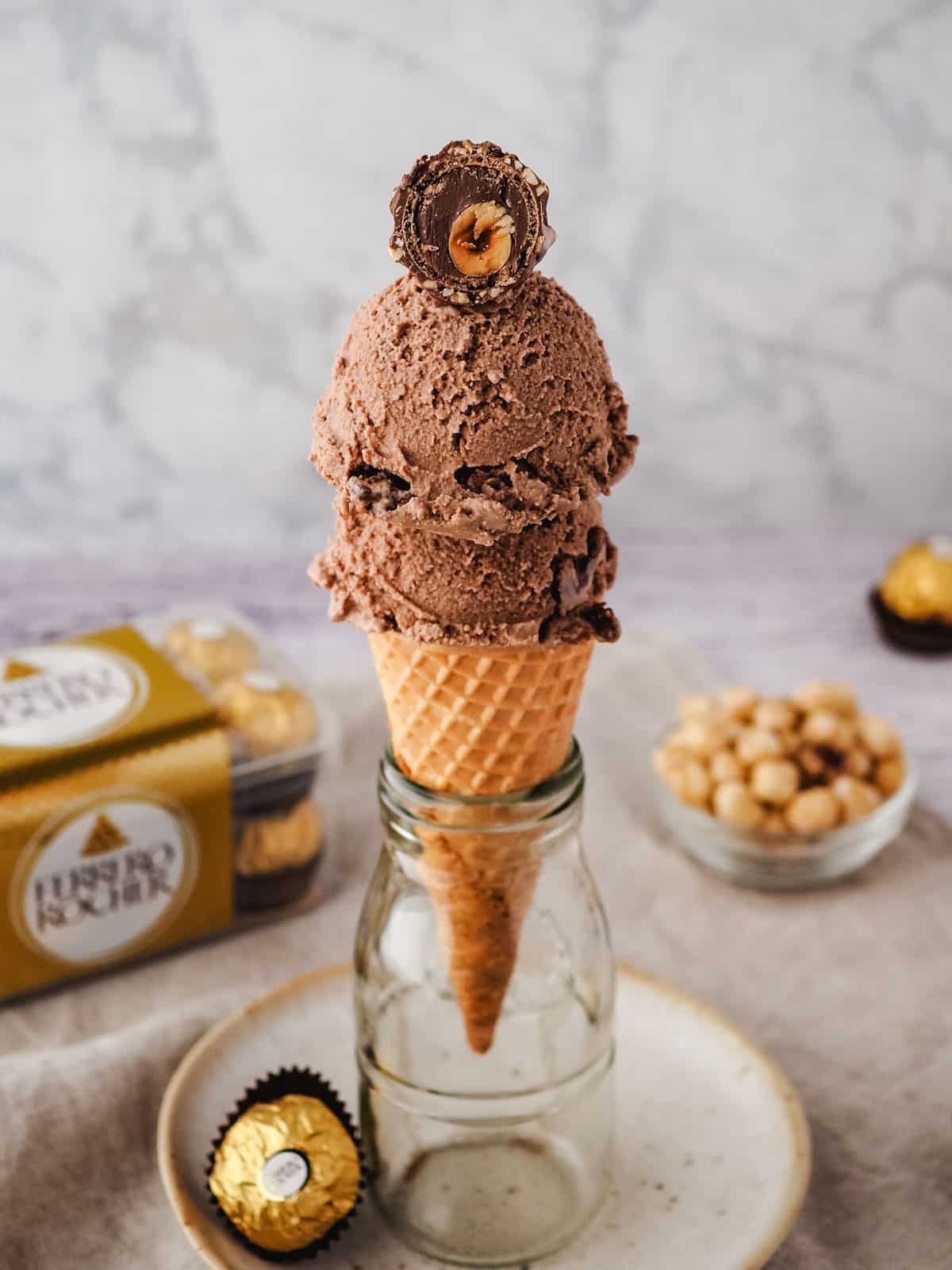 Scoops of Ferrero Rocher ice cream in a cone with a cut-open Ferrero on top, with Ferrero Rocher in the background.