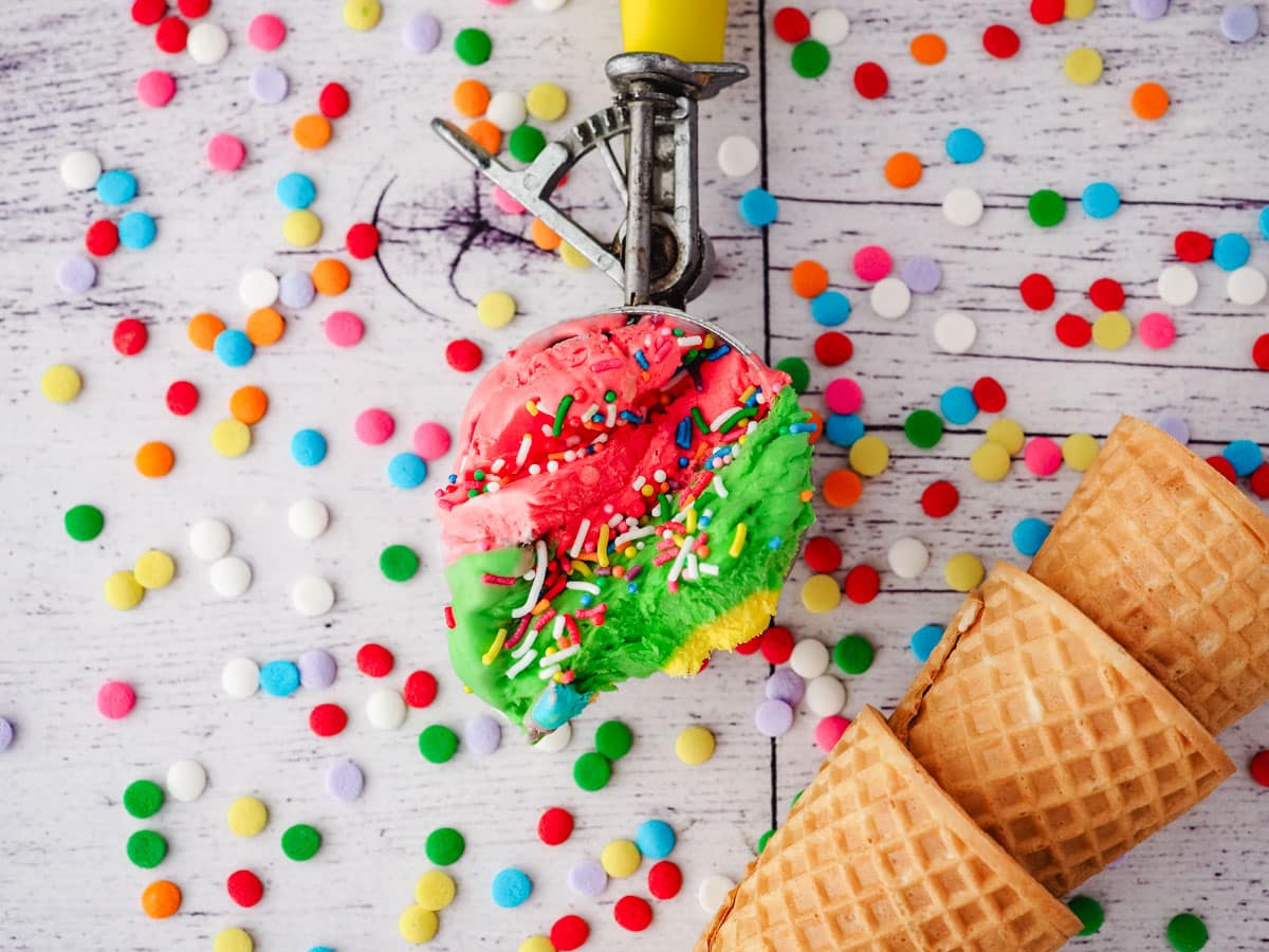 Rainbow ice cream in a in an ice cream scoop, with rainbow sprinkles and ice cream cones on the side.