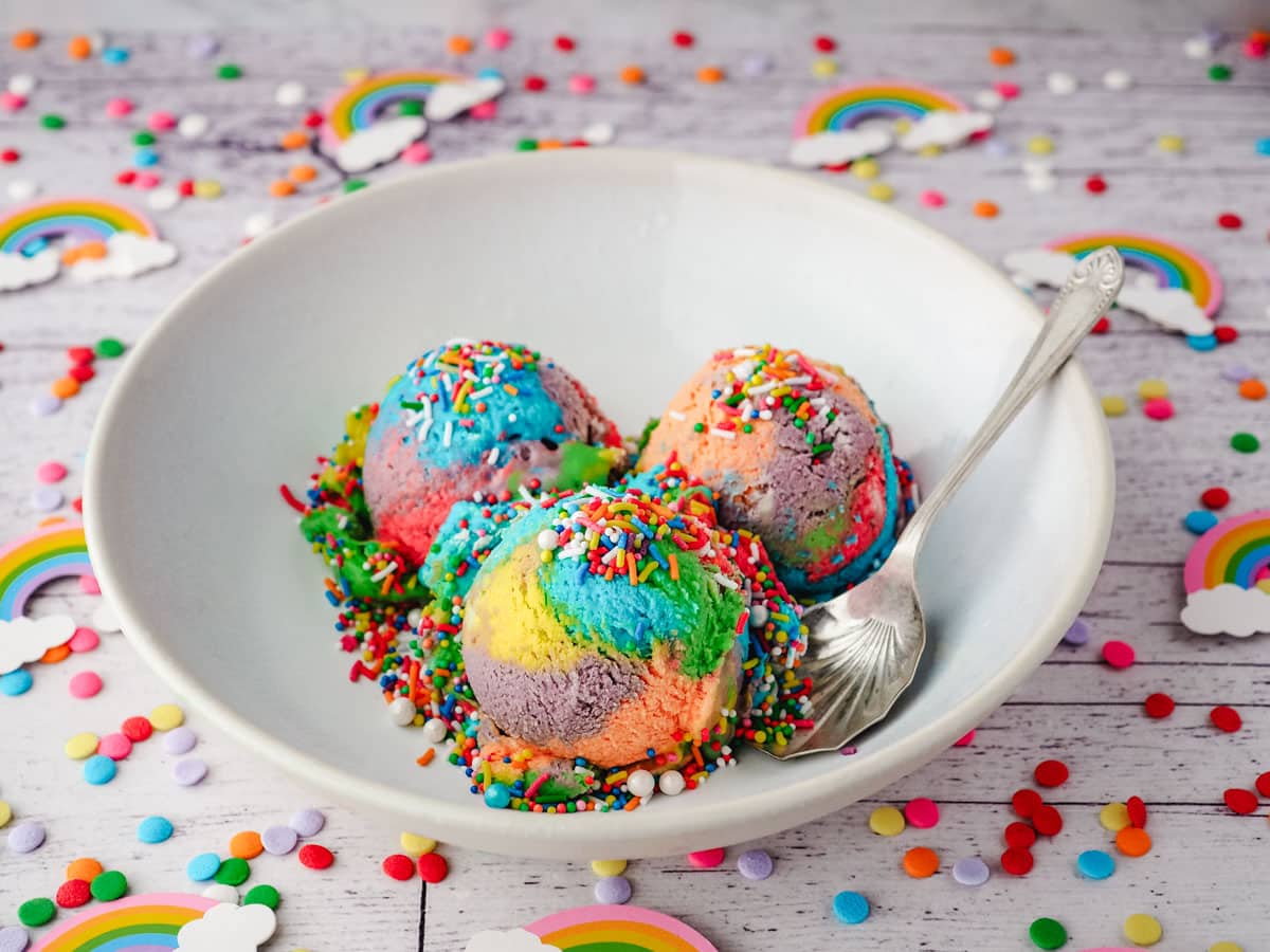 Three scoops of rainbow ice cream with sprinkles in a bowl, with a spoon on the side.