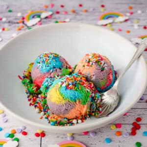 Three scoops of rainbow ice cream with sprinkles in a bowl, with a spoon on the side.