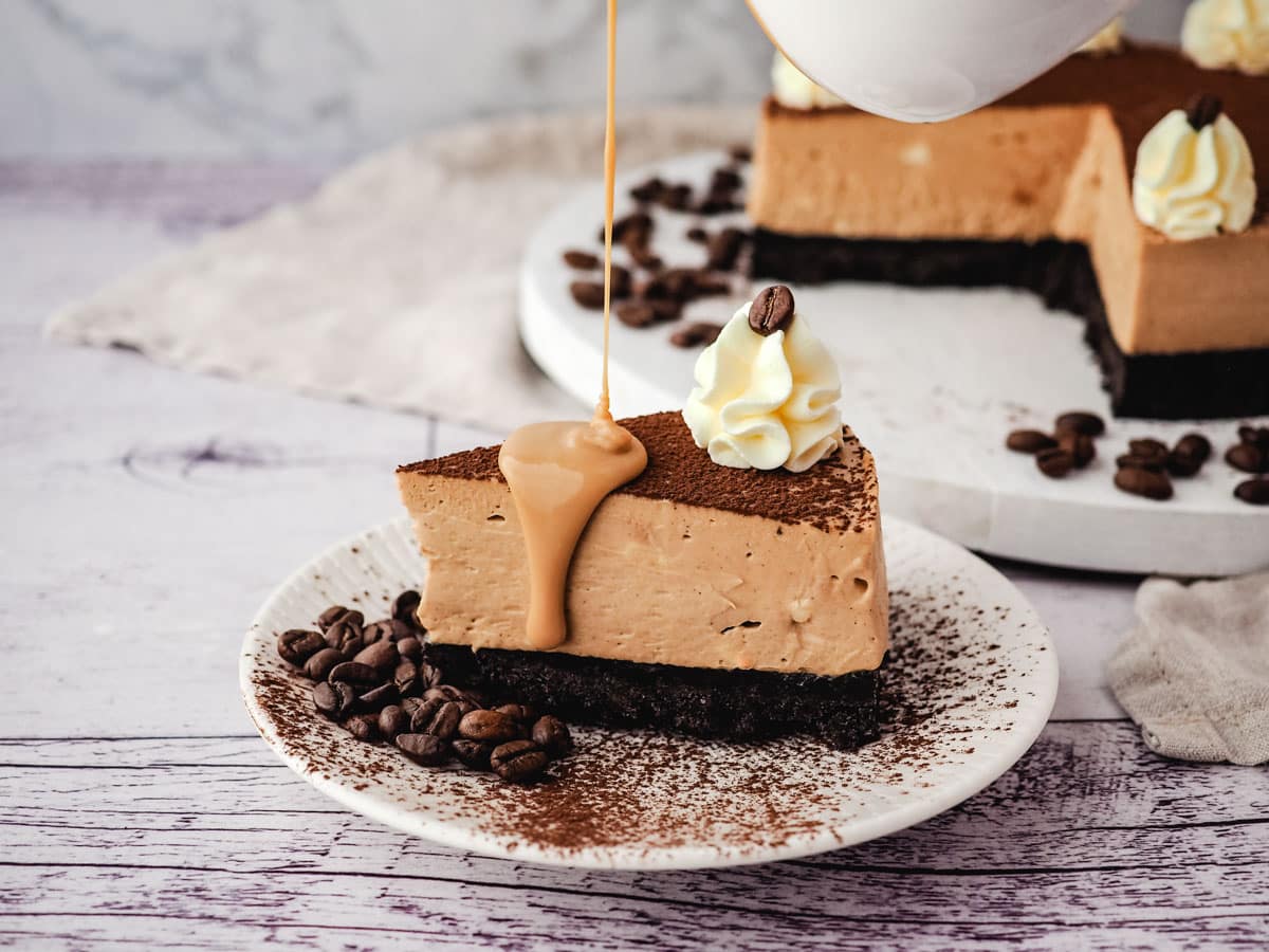 A slice of coffee cheesecake with coffee sauce being drizzled onto it, with rest of cheesecake in the background.