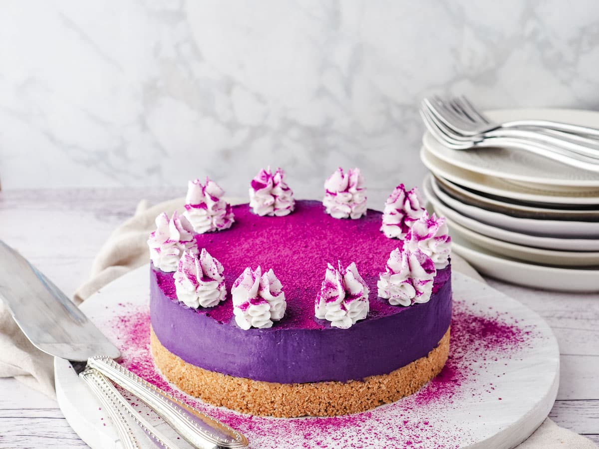 Ube cheesecake with coconut on a serving board, decorated with purple yam powder, with serving utensils and a stack of plates and forks.